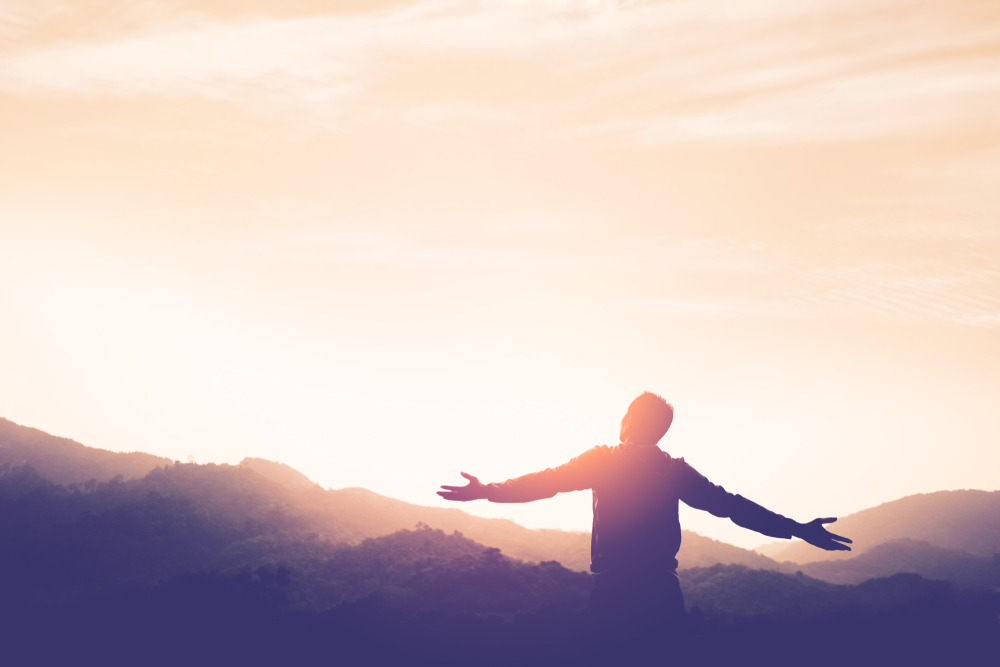 Osoba stojaca na kopci s rozpaženými rukami, obrátená k vychádzajúcemu slnku, symbolizuje nový začiatok a slobodu.
