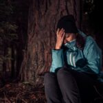 Žena v bundy sedí pod stromom, drží sa za hlavu a zostáva v najťažšej situácii v horách, kde nie je signál a pomoc.