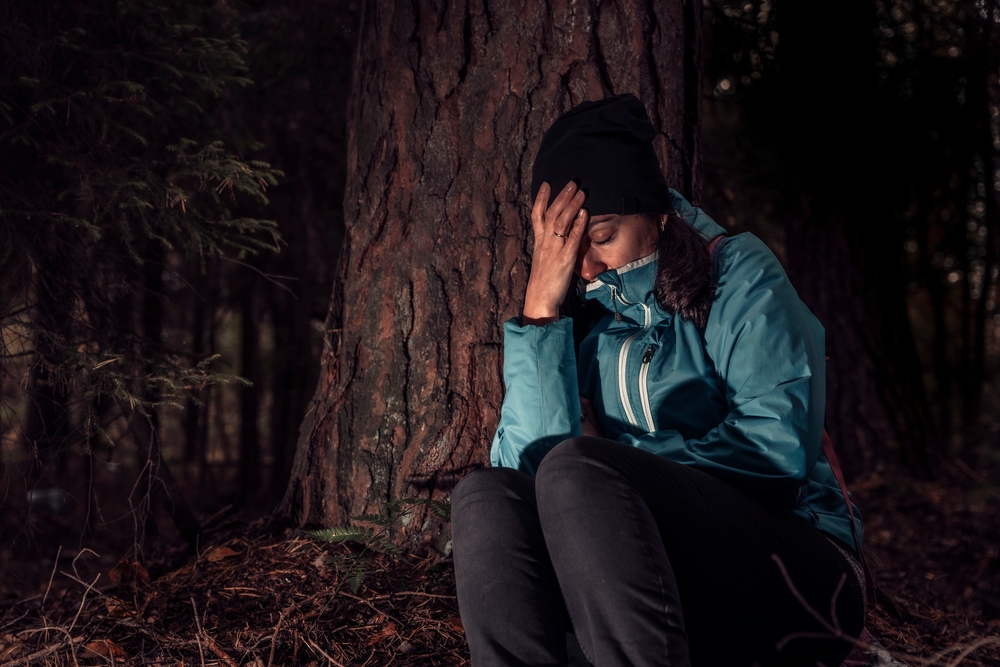 Žena v bundy sedí pod stromom, drží sa za hlavu a zostáva v najťažšej situácii v horách, kde nie je signál a pomoc.